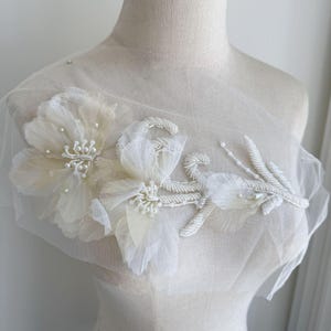 Può includere: Applicazione floreale avorio con due grandi fiori in tessuto e un design a stelo perlato. I fiori hanno petali delicati e sono adornati con piccole perle. L'applicazione è su una rete trasparente bianco sporco.