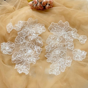 Może przedstawiać: Dwa białe aplikacje z koronki kwiatowej z perłami i cekinami. Aplikacje znajdują się na beżowym tle z tiulu.