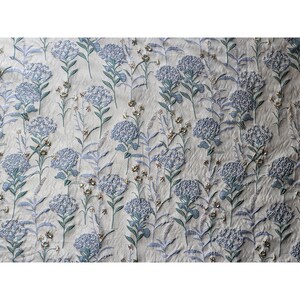 May include: A white fabric with a blue and green floral pattern. The fabric has a textured, embroidered look.