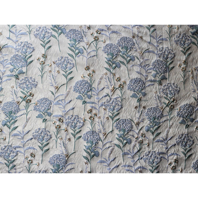 May include: A white fabric with a blue and green floral pattern. The fabric has a textured, embroidered look.