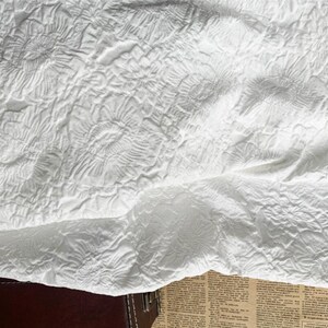 May include: Close-up of a white, textured fabric with a floral pattern. The fabric is folded, revealing layers and shadows. The design features raised, swirling floral motifs, creating a tactile and visually interesting surface. The overall effect is soft and elegant.