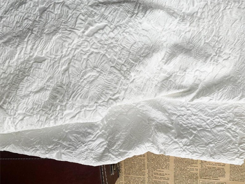 May include: Close-up of a white, textured fabric with a floral pattern. The fabric is folded, revealing layers and shadows. The design features raised, swirling floral motifs, creating a tactile and visually interesting surface. The overall effect is soft and elegant.