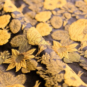 May include: Close-up of a black fabric with a gold floral pattern. The fabric has a textured, embroidered look. The flowers are stylized and resemble sunflowers, roses, and other blooms.