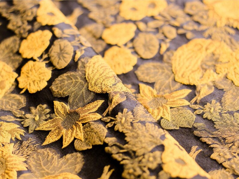 May include: Close-up of a black fabric with a gold floral pattern. The fabric has a textured, embroidered look. The flowers are stylized and resemble sunflowers, roses, and other blooms.