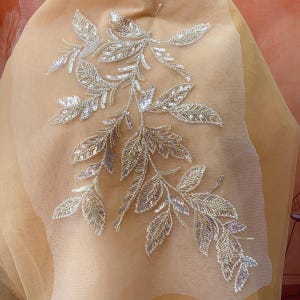 Può includere: Un delicato disegno di foglie dorate e argentate ricamate su un tessuto beige trasparente. Le foglie sono disposte in un motivo fluido, creando un design bellissimo e intricato.