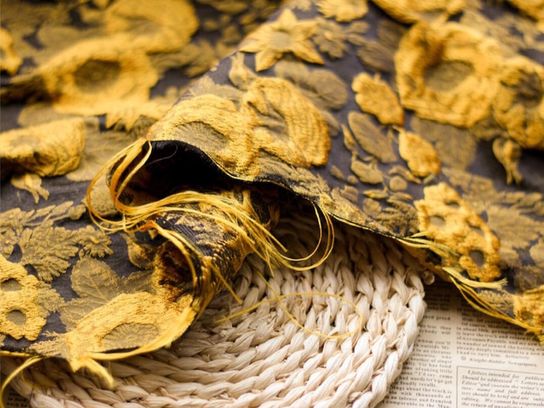 May include: Close-up of a gold and black fabric with a floral pattern. The fabric is folded and draped over a woven basket.