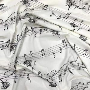 Op de afbeelding: Witte stof met een zwart muzieknoot- en rozenpatroon. De stof is gevouwen en toont het patroon aan beide zijden.