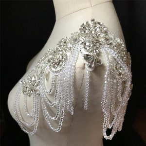 May include: A silver and white beaded shoulder piece with a large crystal embellishment. The piece is designed to be worn on one shoulder and has a cascading effect of beads.