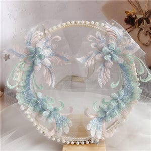 Puede incluir: Una delicada aplicación de encaje floral con ribete de perlas. La aplicación presenta un diseño floral de color azul claro y rosa sobre una tela blanca transparente.