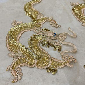 Könnte beinhalten: Gestickte goldene Drachenapplikation mit aufwendigen Details. Der Drache hat goldene Pailletten, hellrosa Stickereien und blaue Edelsteinaugen. Das Design befindet sich auf einem hellbeigen Hintergrund. Für Kleidung oder Bastelprojekte.