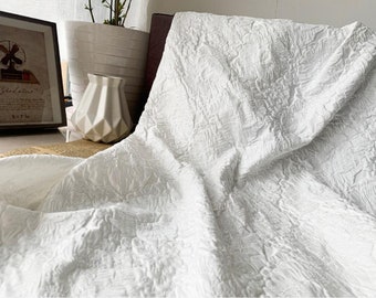 May include: A close-up of a white textured fabric with a raised, floral-like pattern. The fabric is draped, creating soft folds and shadows. The image also shows a framed picture, a white vase, and a potted plant.