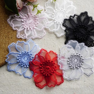 May include: Six floral appliques in a range of colours: pale pink, white, black, light blue, red, and silver. Each applique has a detailed, layered design with a central, textured element. The appliques are made of a sheer, delicate fabric.