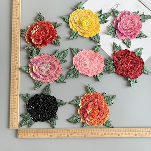 Op de afbeelding: Een verzameling bloemenapplicaties met pailletten in verschillende kleuren, waaronder rood, geel, roze en zwart. Elke bloem heeft groene bladeraccenten. De applicaties zijn gerangschikt op een grijze ondergrond.