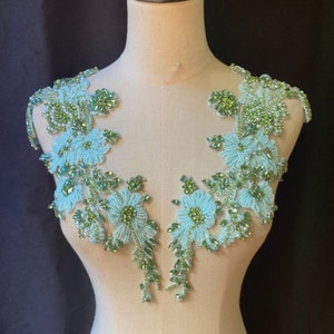 Puede incluir: Dos aplicaciones florales de color azul claro con abalorios verdes y blancos y pedrería. Las aplicaciones están en un maniquí blanco.