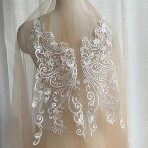 May include: White lace applique with a floral design. The lace is delicate and intricate, with a variety of floral motifs. The applique is perfect for adding a touch of elegance to any garment.