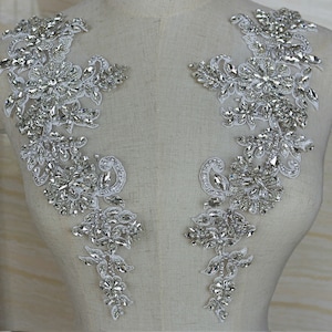 May include: Two silver rhinestone applique embellishments with a floral design. The embellishments are on a white fabric background.