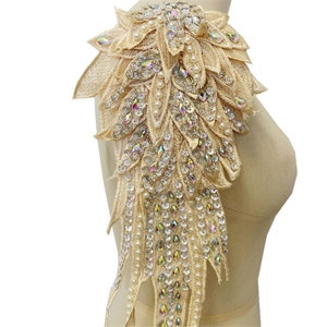 May include: A beige fabric applique with a wing shape, decorated with iridescent rhinestones and pearl beads.