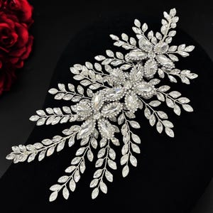 Puede incluir: Un peine de pelo de novia plateado con detalles de cristal transparente y forma de hojas. El peine presenta un diseño floral con múltiples racimos de cristal y delicadas ramitas de hojas, ideal para bodas u ocasiones especiales.