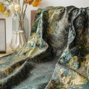 May include: A draped fabric with a mottled pattern in shades of green, gold, and black. The fabric is textured and appears to be soft. A framed picture and a vase with flowers are in the background.