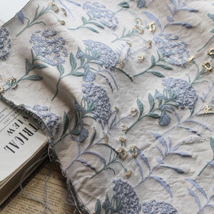 May include: A close-up of a white fabric with a blue and gold floral pattern. The fabric has a textured surface and appears to be woven. The fabric is folded and draped over a book.