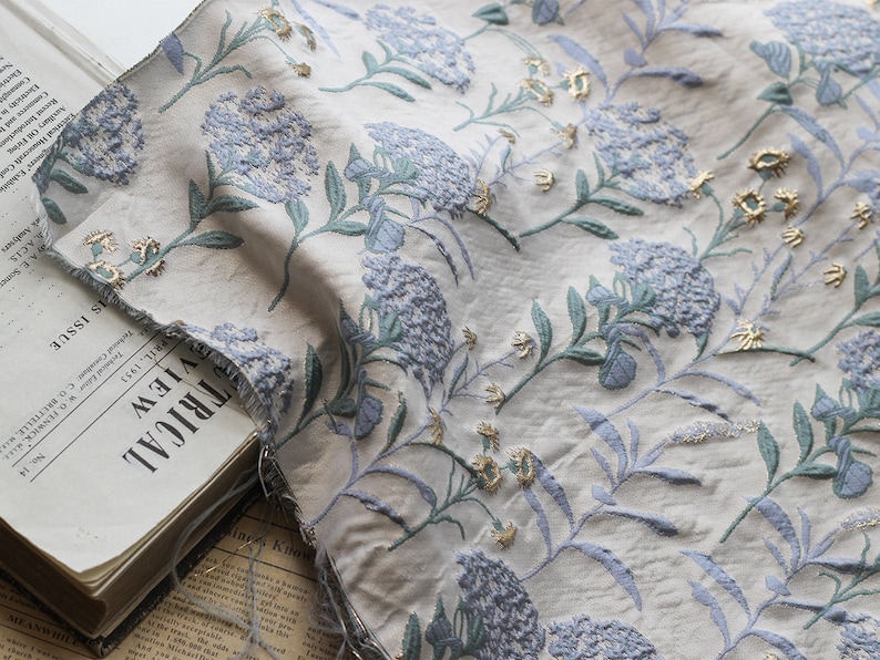 May include: A close-up of a white fabric with a blue and gold floral pattern. The fabric has a textured surface and appears to be woven. The fabric is folded and draped over a book.