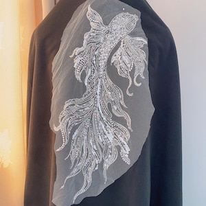 May include: A white fish-shaped applique with silver sequins and beads on a sheer white fabric. The applique is ideal for adding a touch of sparkle to clothing, bags, or home decor.