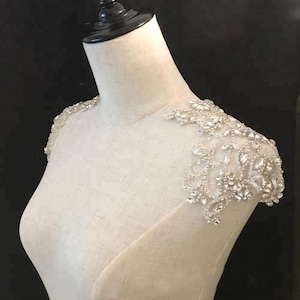 Può includere: Un paio di spalline con cristalli per un abito da sposa o altri abiti formali. Le spalline sono realizzate in cristalli trasparenti e hanno un design floreale delicato.