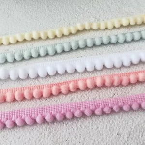 May include: Five spools of pom-pom trim in various pastel colours. The colours are light yellow, light blue, white, light pink, and pink.