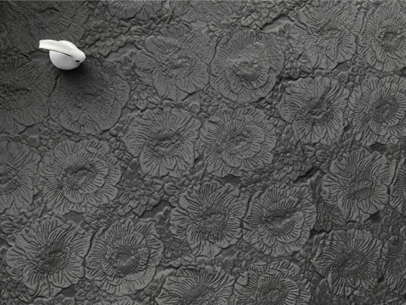 May include: A close-up shot of a dark gray, textured surface with a repeating floral pattern. A small, white, curved object is positioned in the upper left corner. The surface has a raised, embossed design of stylized flowers.
