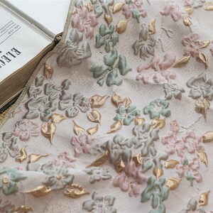 May include: Close-up of a white fabric with a floral pattern. The fabric features raised, embroidered flowers in shades of pink, green, and gold.