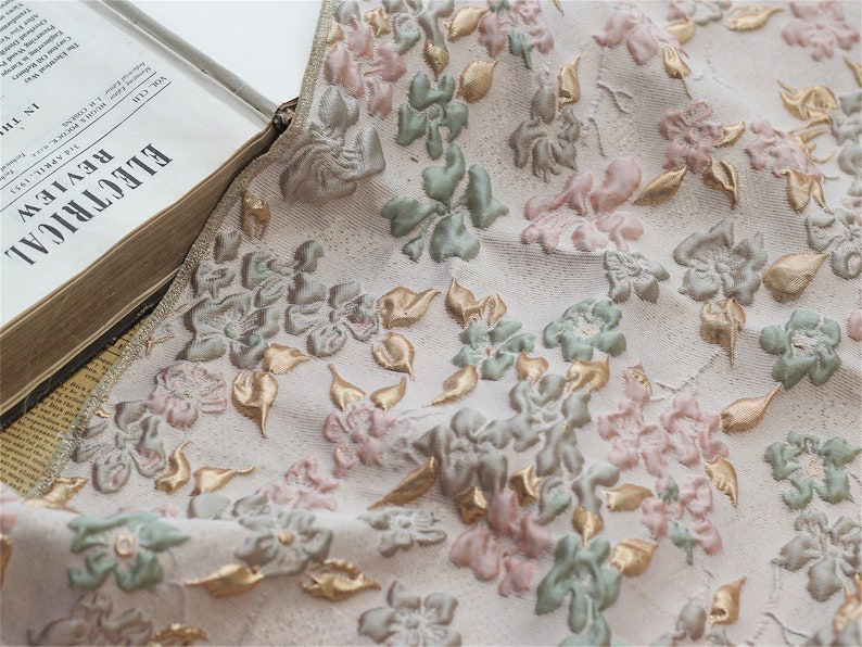 May include: Close-up of a white fabric with a floral pattern. The fabric features raised, embroidered flowers in shades of pink, green, and gold.