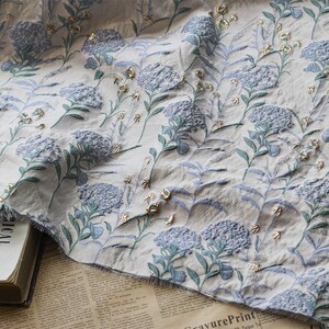 May include: A white fabric with a blue and green floral pattern. The flowers are embossed and have a gold metallic sheen.