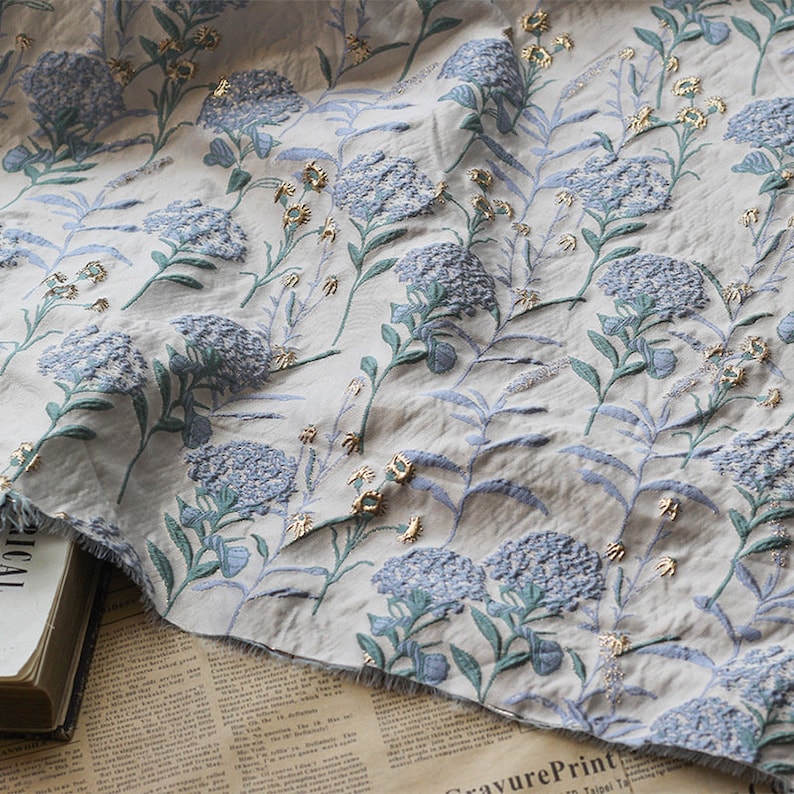May include: A white fabric with a blue and green floral pattern. The flowers are embossed and have a gold metallic sheen.