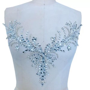May include: A silver rhinestone applique with a V-shaped design. The applique is made of small, round rhinestones that are arranged in a floral pattern.
