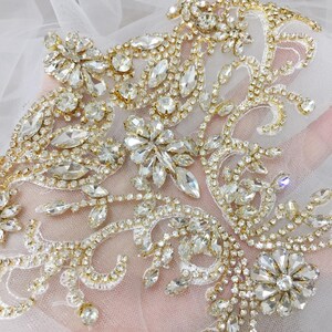 May include: Close-up of a decorative embellishment featuring clear, sparkling rhinestones and gold-colored metal. The design includes floral and leaf patterns, with various sizes and shapes of rhinestones. The embellishment is likely for bridal or formal wear.