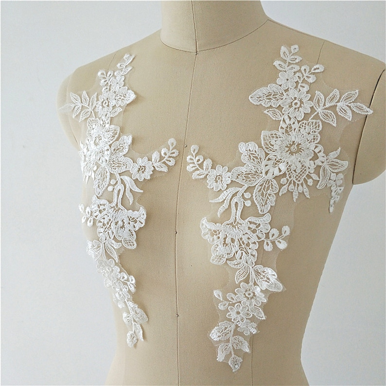 May include: Two white floral lace appliques on a white mannequin. The appliques are made of delicate lace with a floral design. They are perfect for adding a touch of elegance to any garment.
