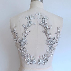 May include: A pair of silver rhinestone applique embellishments for clothing. The embellishments are shaped like floral designs with a delicate, intricate pattern. They are perfect for adding a touch of sparkle and elegance to any garment.