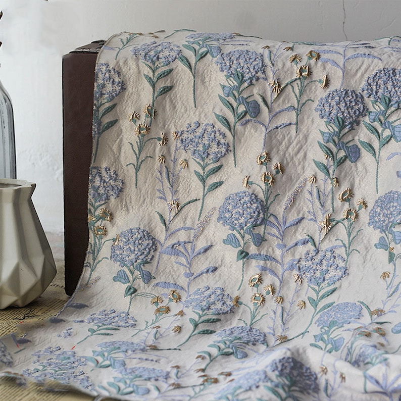 May include: A white fabric with a blue and gold floral pattern. The fabric is textured and has a subtle sheen. The flowers are embroidered with small beads.