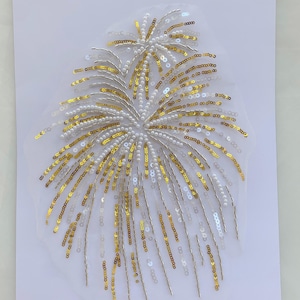 Può includere: Un'applicazione decorativa con un design ispirato ai fuochi d'artificio, realizzata con paillettes dorate, perline argentate e piccole perle bianche. L'ornamento è disposto a raggiera su un supporto in rete bianca semitrasparente.