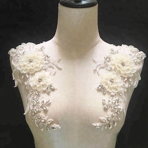 Puede incluir: Dos apliques florales blancos con pedrería y detalles de perlas. Los apliques están en un busto de maniquí blanco.