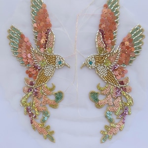 May include: Two symmetrical bird-shaped appliqués featuring intricate beadwork and sequins in shades of peach, gold, light green, and purple. The birds are depicted in flight with outstretched wings and long, flowing tail feathers on a sheer fabric.