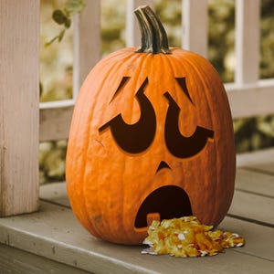 May include: A carved pumpkin with a distressed facial expression, with its mouth open and a pile of orange candy spilling out.