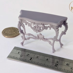 May include: A small, intricately carved, light purple console table with a rectangular top. The table is about 5 inches long and is shown next to a ruler.