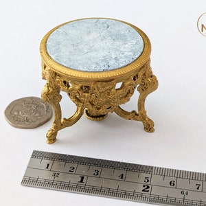 May include: A small, gold-coloured, decorative table with a round, light blue marble top. The table has intricate, ornate details and three legs. A ruler is in the foreground for scale.