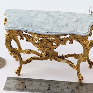 May include: A small, ornate gold console table with a light blue marble top. The table has intricate carvings and a delicate design. It measures approximately 25 cm long.