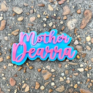 Op de afbeelding: Een roze en blauw harsbord met de tekst "Mother Dearra" in een cursief schrift.