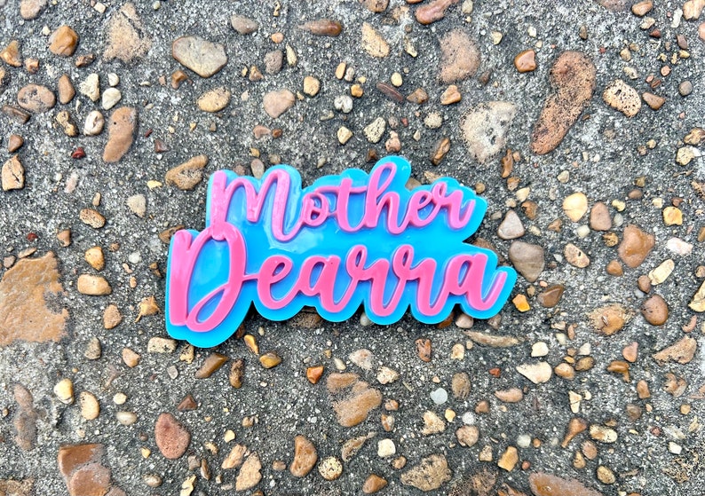 Op de afbeelding: Een roze en blauw harsbord met de tekst "Mother Dearra" in een cursief schrift.