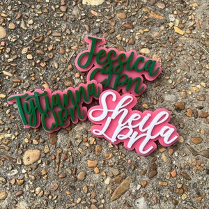Op de afbeelding: Drie gepersonaliseerde naamstickers met de namen "Jessica", "Tytiana" en "Sheila" in cursief. De namen zijn uit groen en wit acryl geknipt en zijn op roze acryl gelegd.