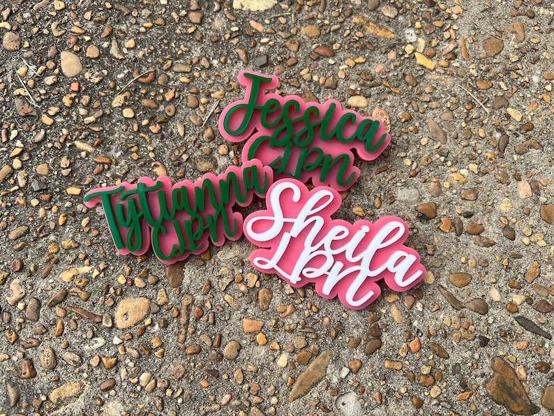 Op de afbeelding: Drie gepersonaliseerde naamstickers met de namen "Jessica", "Tytiana" en "Sheila" in cursief. De namen zijn uit groen en wit acryl geknipt en zijn op roze acryl gelegd.