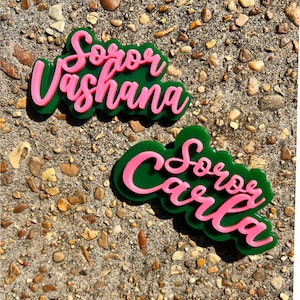 May include: Two layered acrylic name tags. The bottom layer is green, and the top layer is pink. The top tag says "Soror Vashana" in cursive. The bottom tag says "Soror Carla" in cursive.